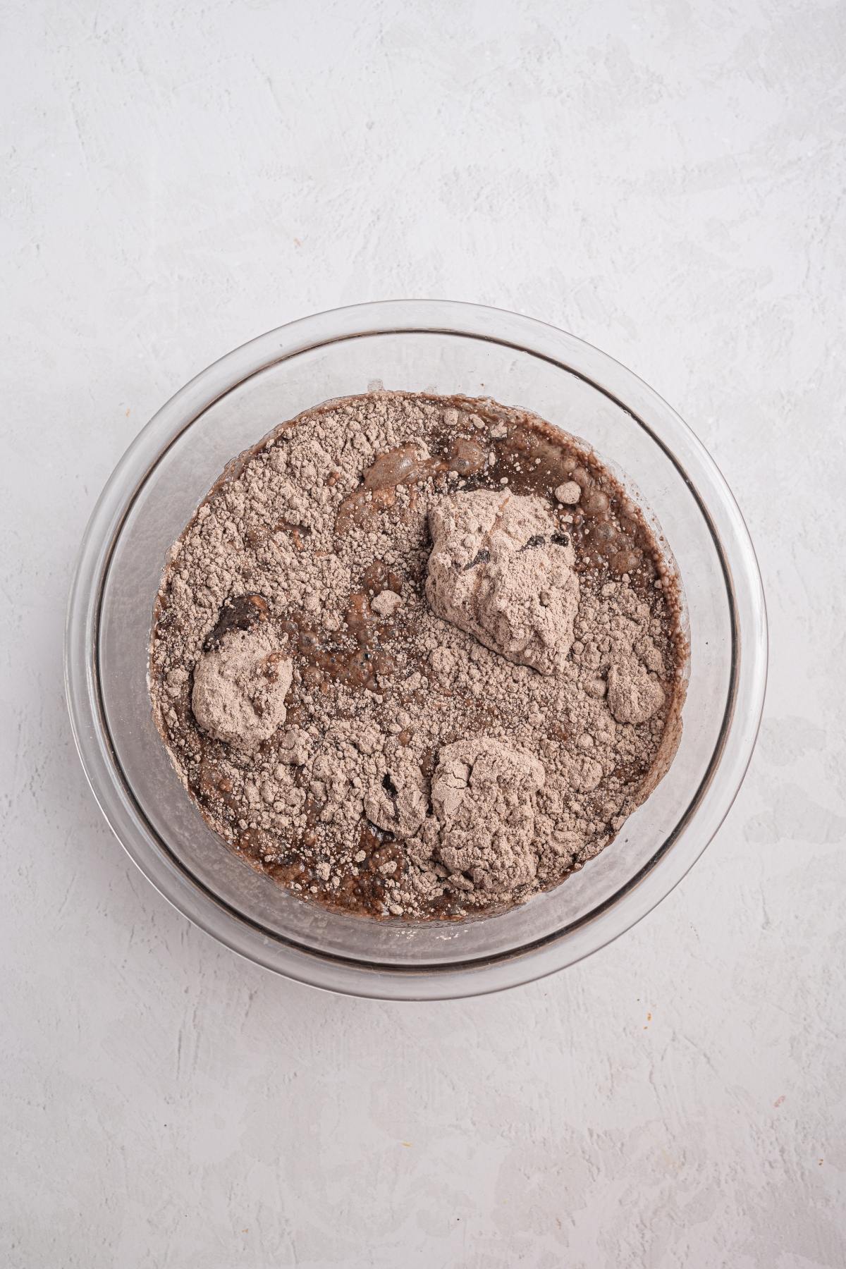 A glass bowl with dry chocolate cake mix and wet ingredients, not yet mixed, on a light surface.