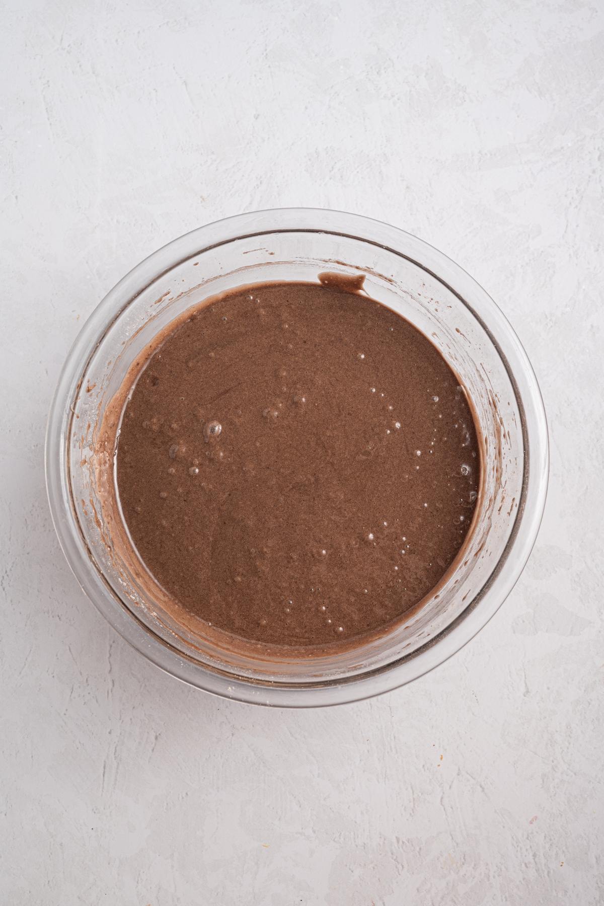 A glass bowl filled with chocolate cake batter on a light-colored surface.