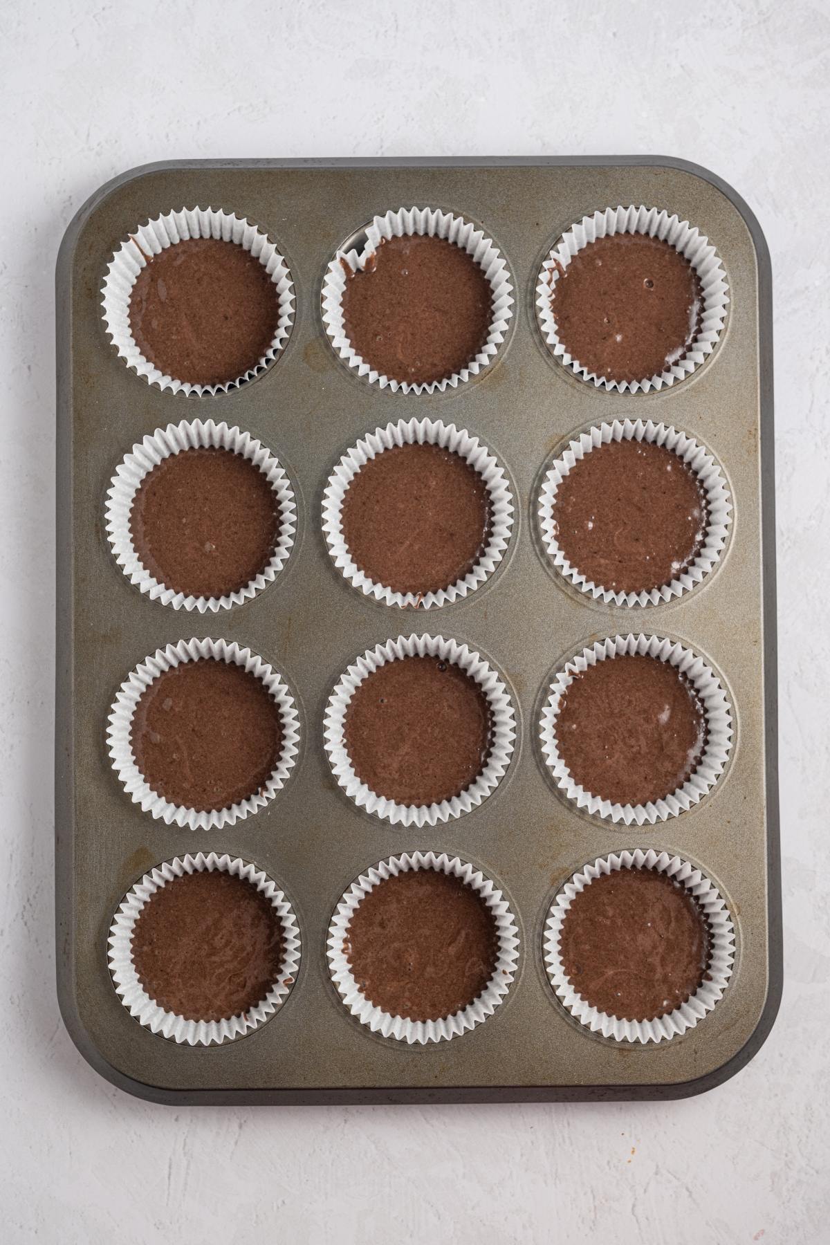 Muffin tin with twelve paper liners filled with chocolate cupcake batter, ready to be baked.