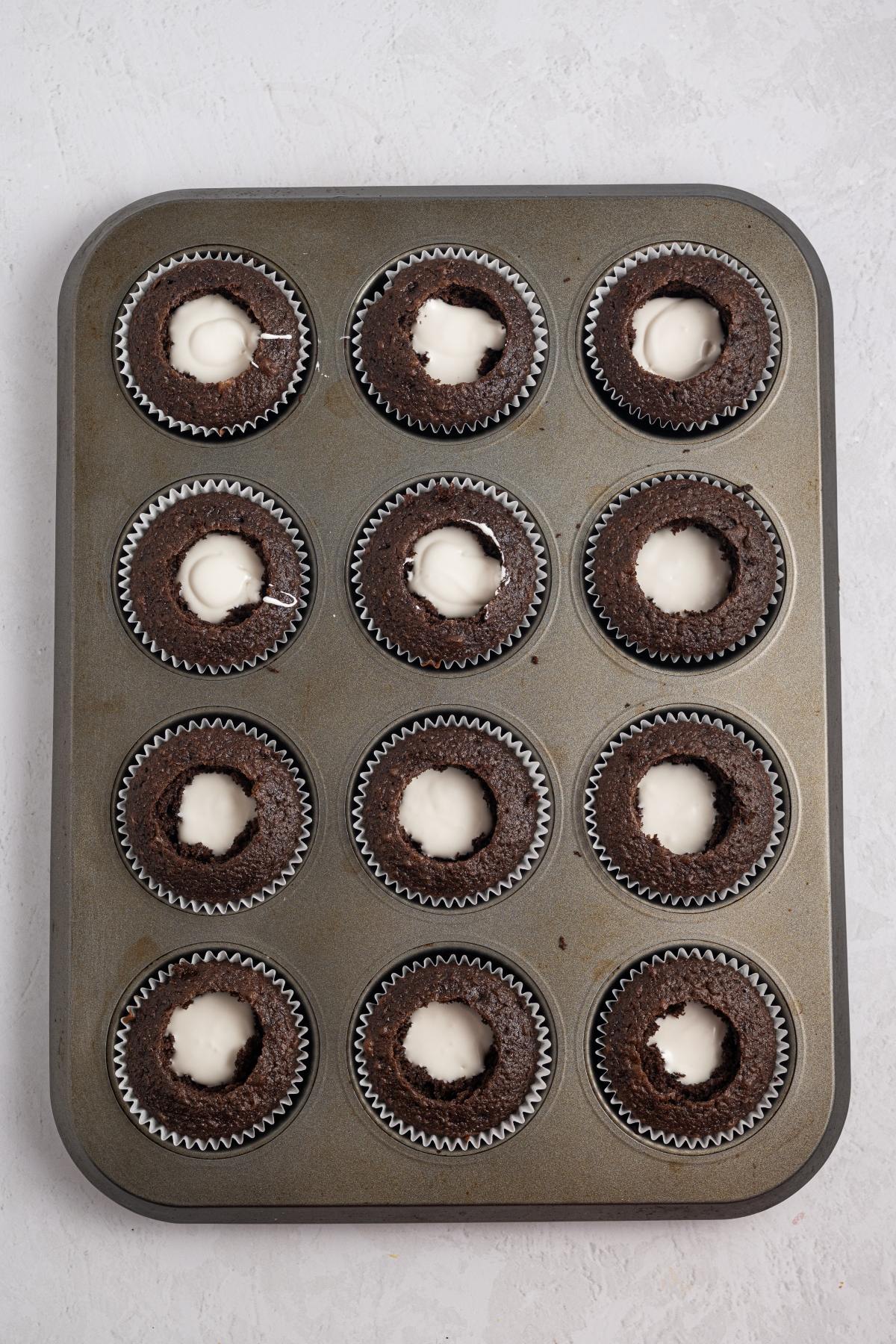 A muffin tin with 12 chocolate cupcakes, each with a white cream filling in the center.