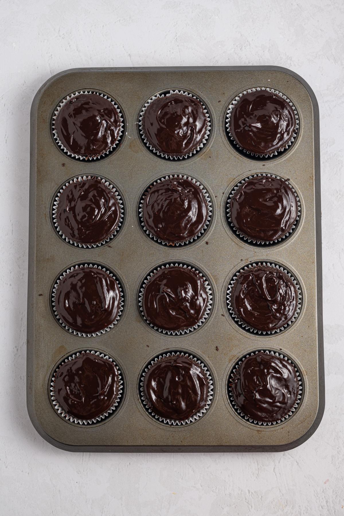 A muffin tin with 12 cupcake liners filled with chocolate batter on a light surface.