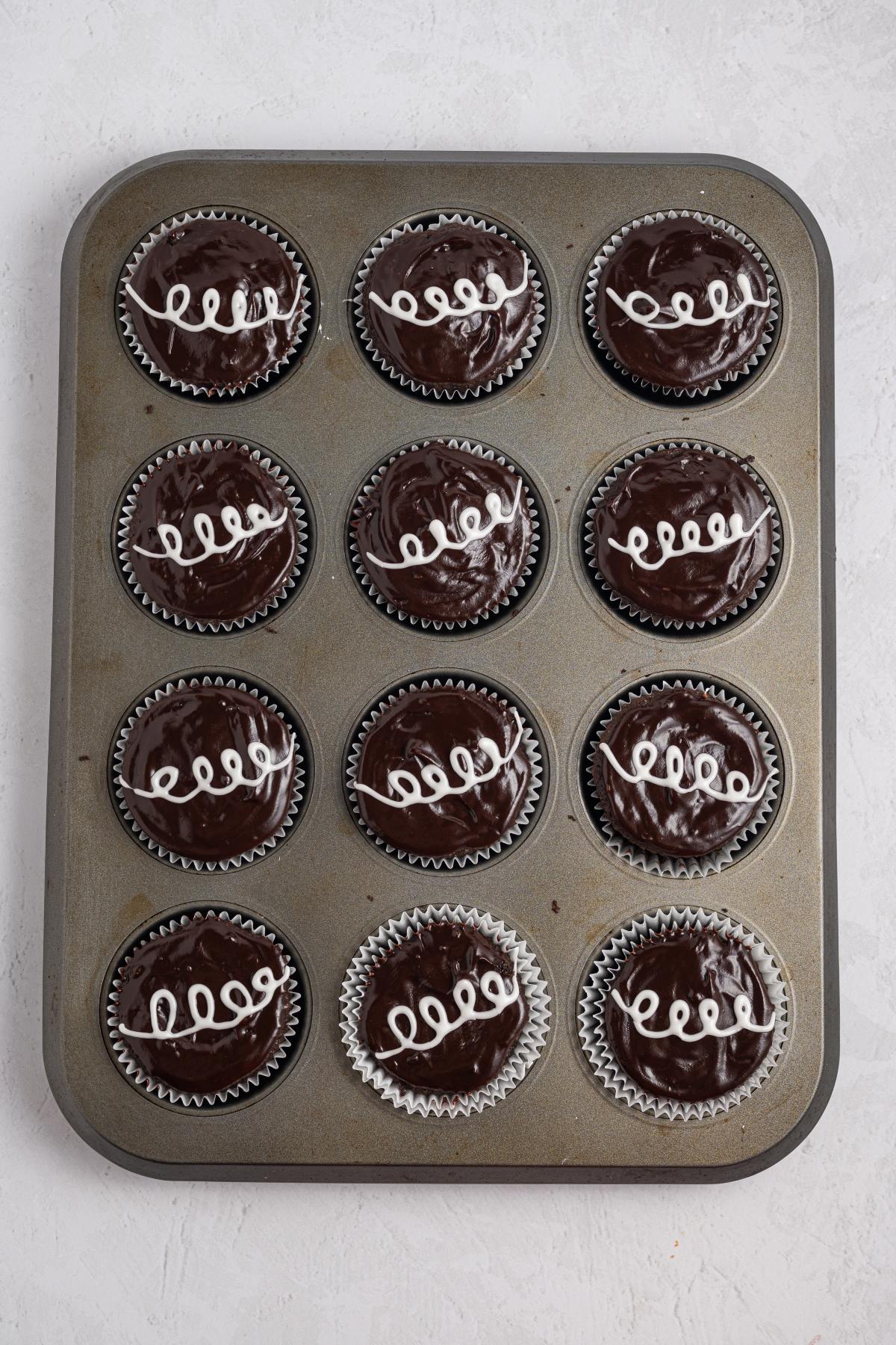 A muffin tin with twelve chocolate cupcakes, each topped with a white icing swirl.