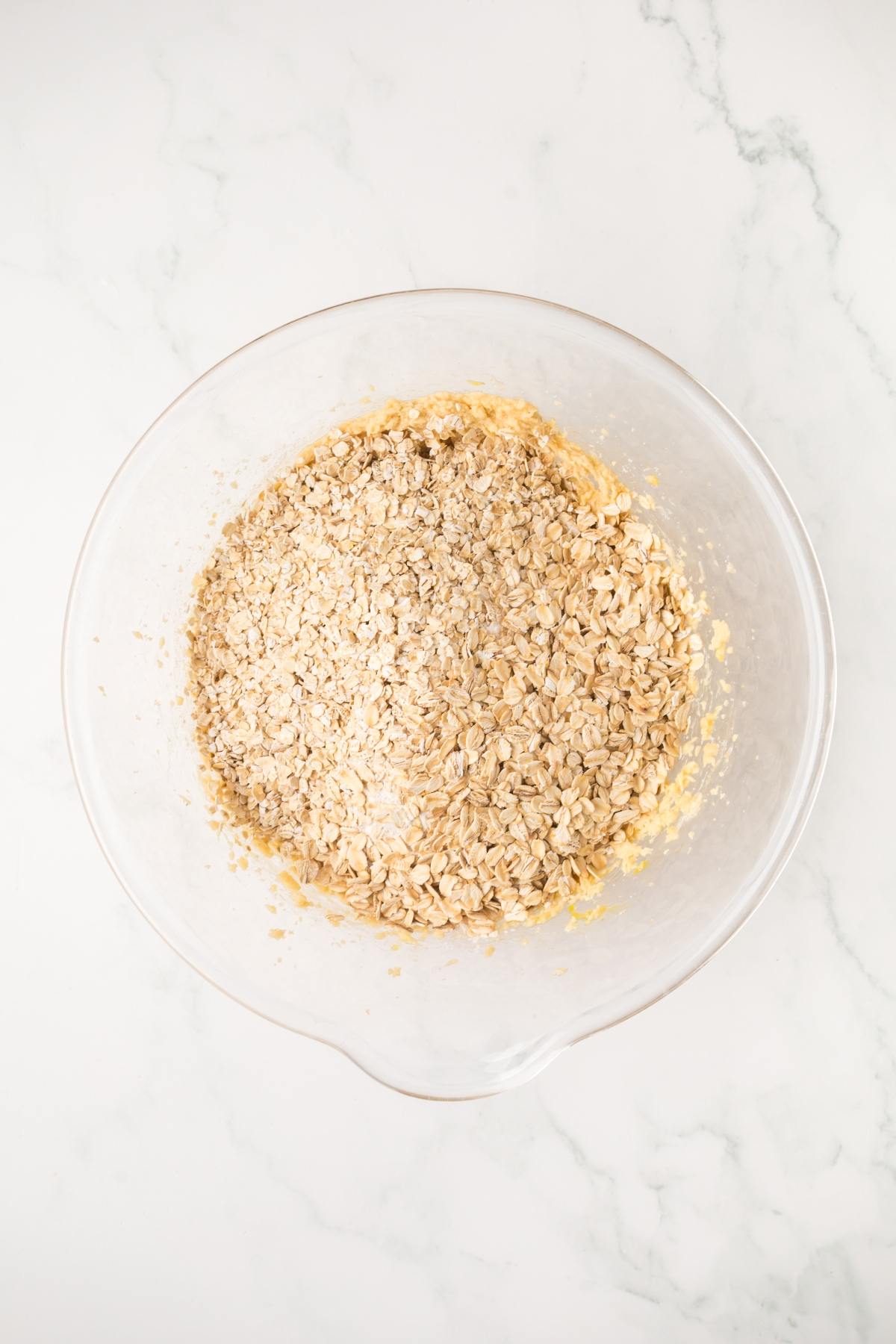 Clear bowl with oats and dough mixture on a white marble surface, ready for mixing.