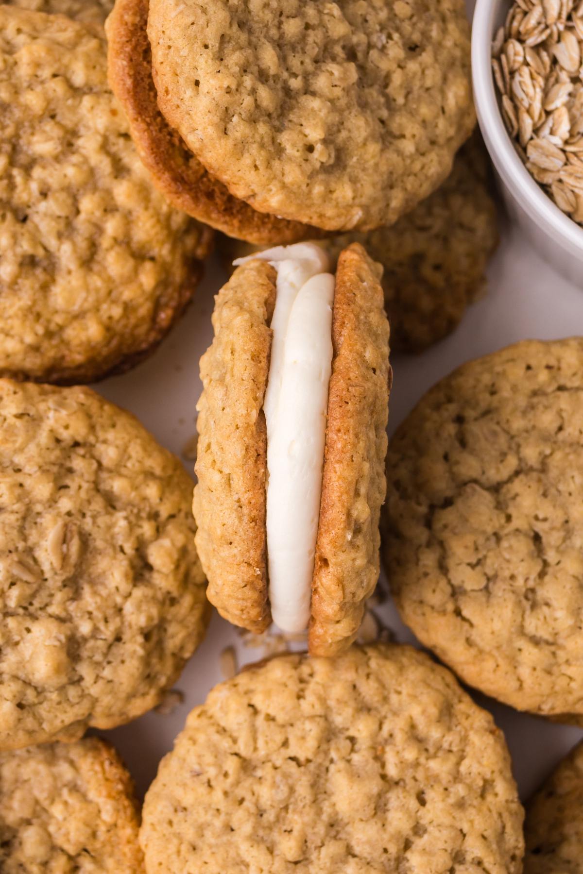 Oatmeal cream pies with a creamy filling, surrounded by several plain oatmeal cookies and a bowl of oats.