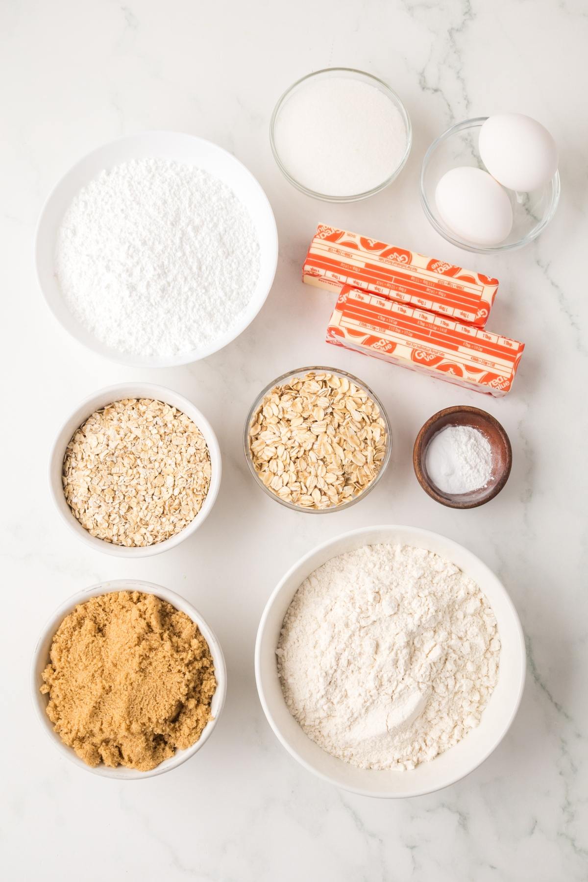 Baking ingredients including flour, oats, sugars, butter, eggs, and baking soda arranged on a white surface.