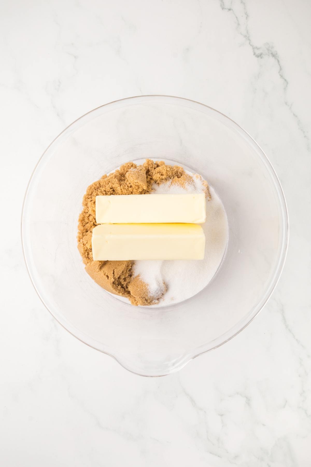 Two sticks of butter, brown sugar, and white sugar in a clear mixing bowl on a marble countertop.