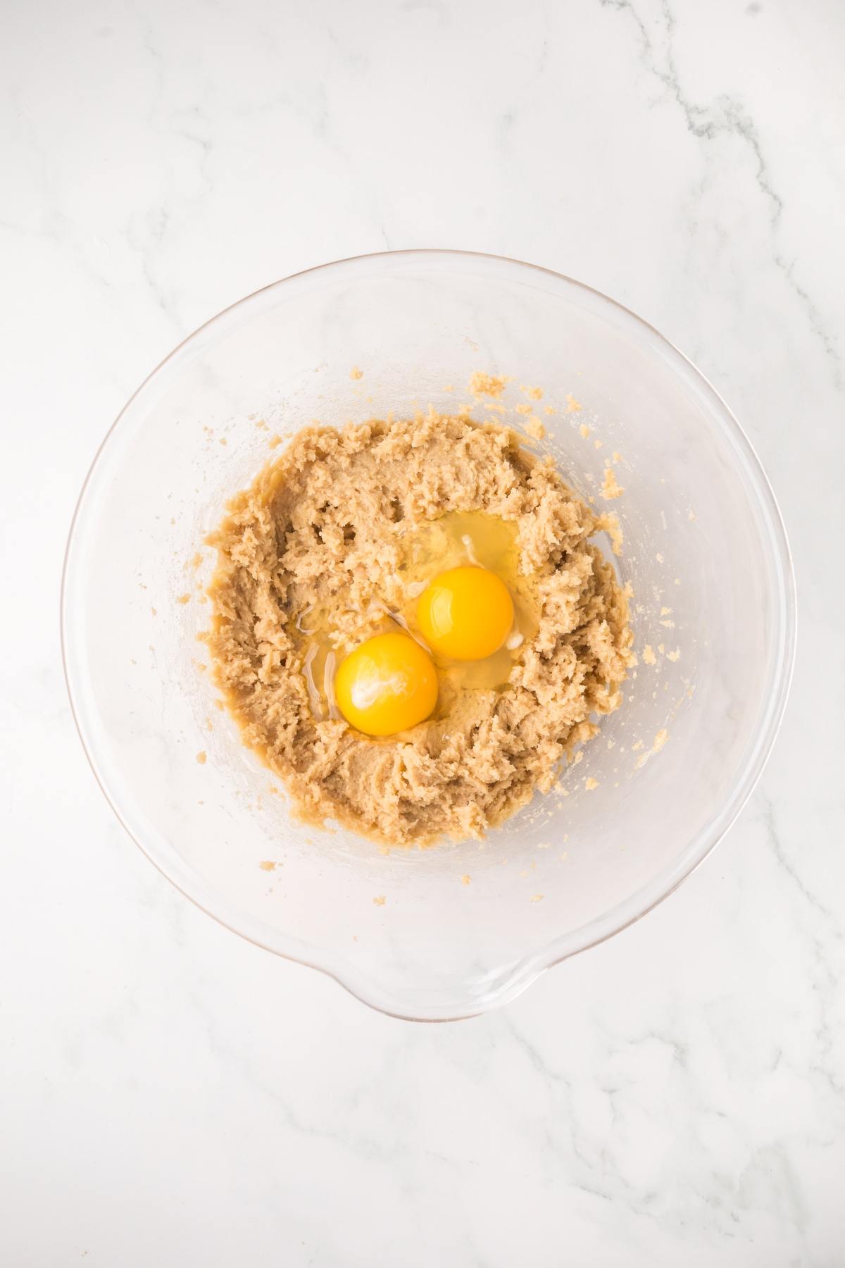 A glass bowl with cookie dough mixture and two raw eggs on a white marble surface.