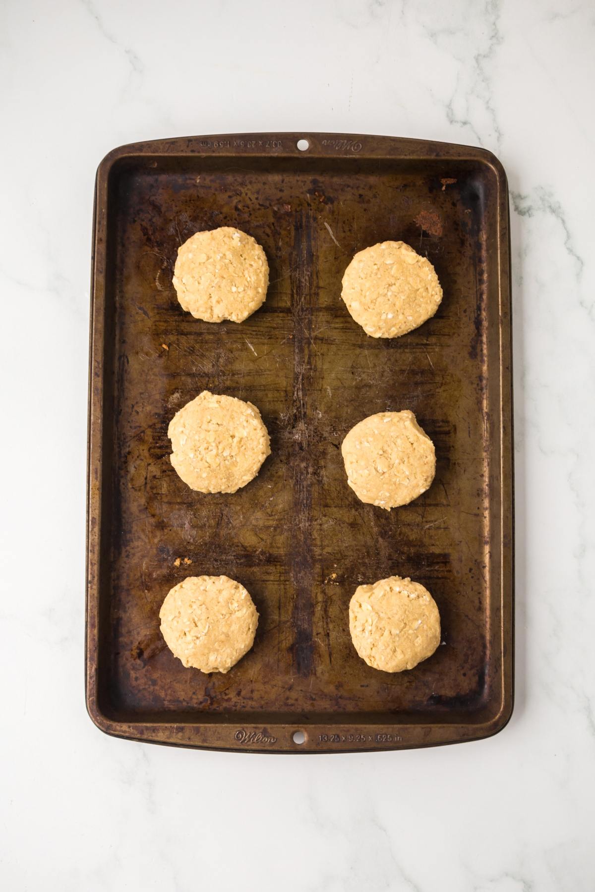Six unbaked cookies are arranged on a worn baking sheet atop a white marble surface.