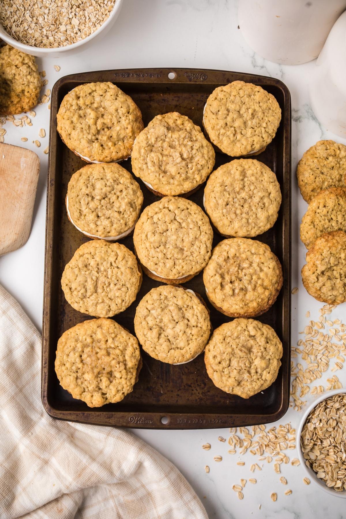 A baking tray with twelve oatmeal oatmeal cream pies, surrounded by scattered oats and extra cookies on a white surface.