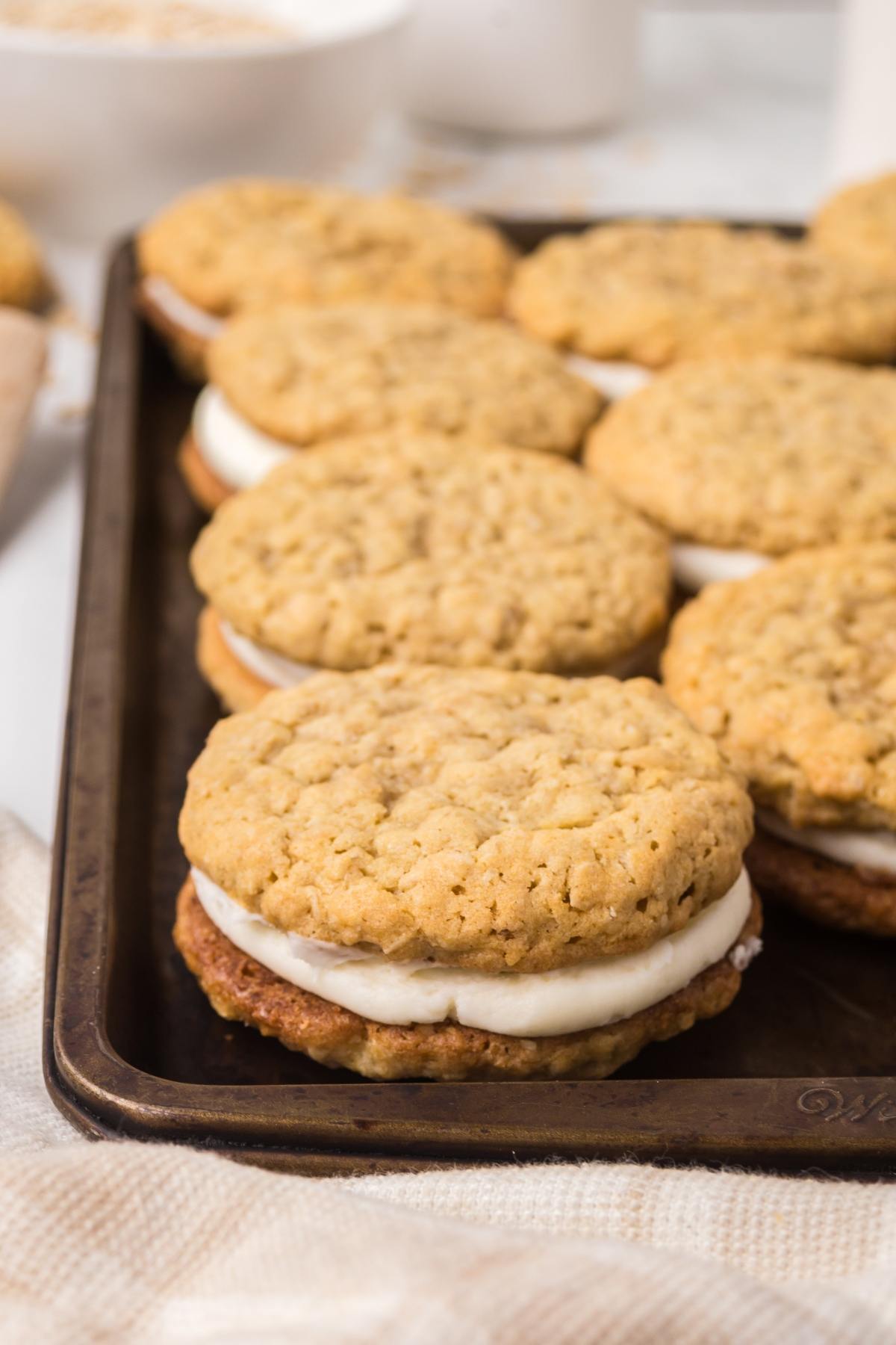 Several oatmeal cream pies with white filling are arranged on a dark baking tray.