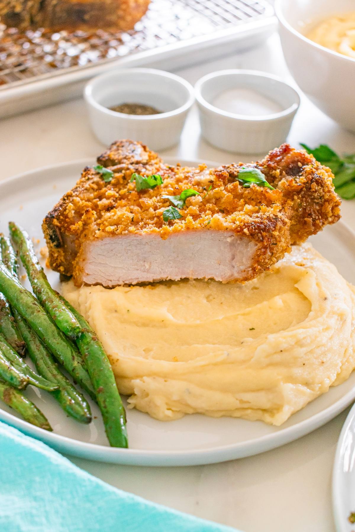Breaded pork chop on mashed potatoes with green beans, garnished with herbs, on a white plate.