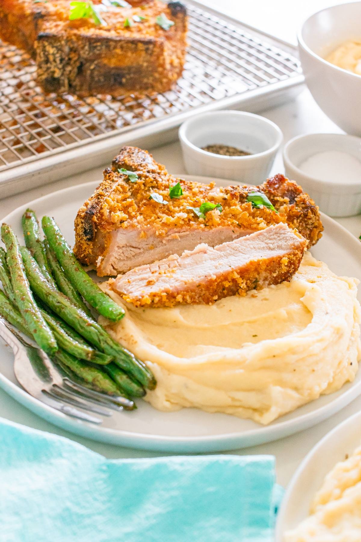 A breaded pork chop with mashed potatoes and green beans on a white plate, garnished with herbs.