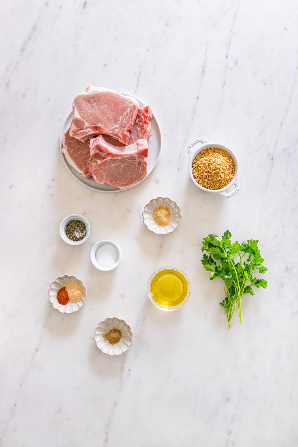 Raw pork chops, rice, fresh cilantro, olive oil, and various spices arranged on a white marble surface.