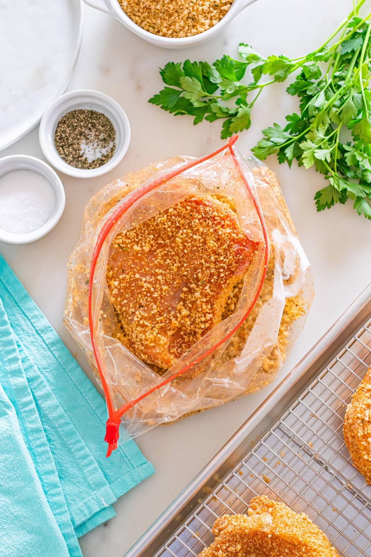 Seasoned breaded pork chops in a zip bag, surrounded by spices, herbs, and a blue napkin on a white counter.