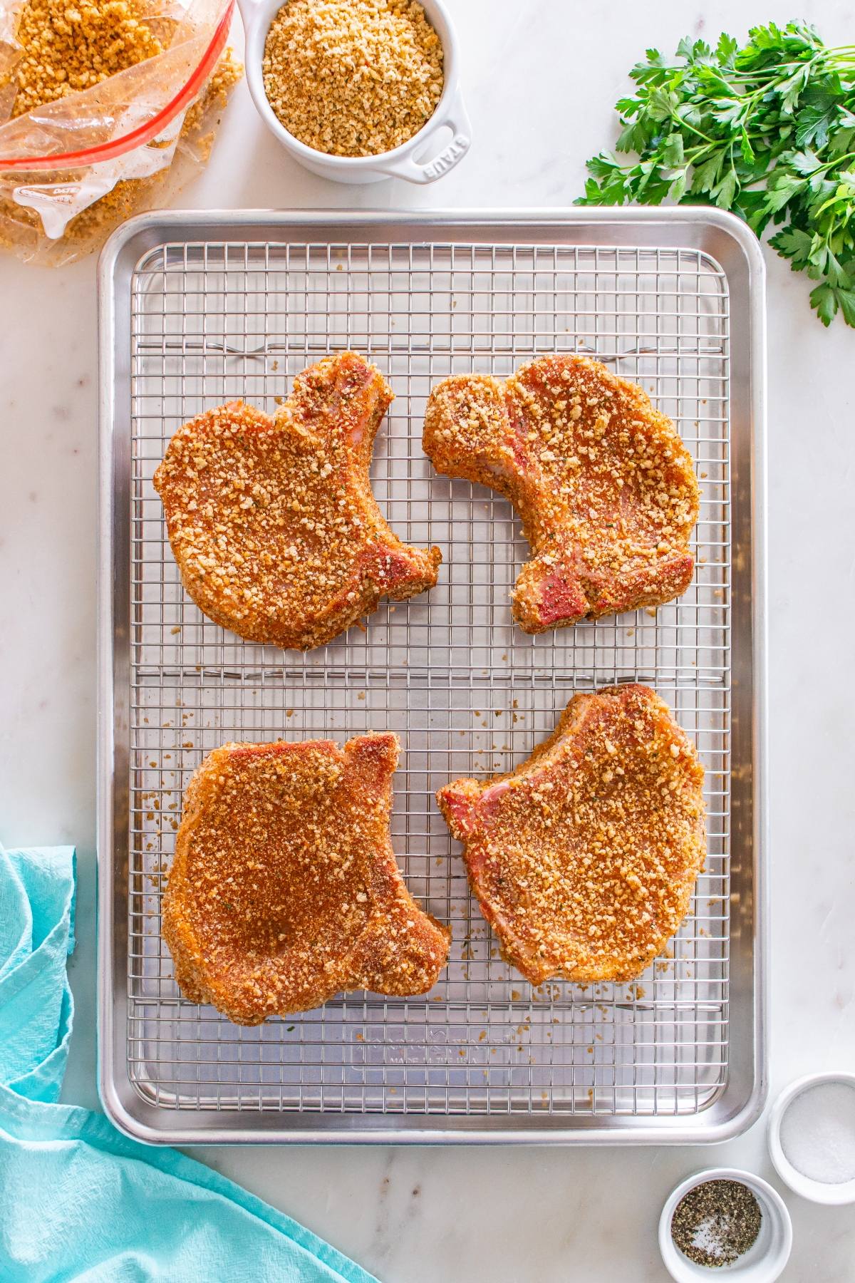 Four breaded pork chops on a wire rack over a baking sheet, with parsley, crumbs, and seasonings nearby.