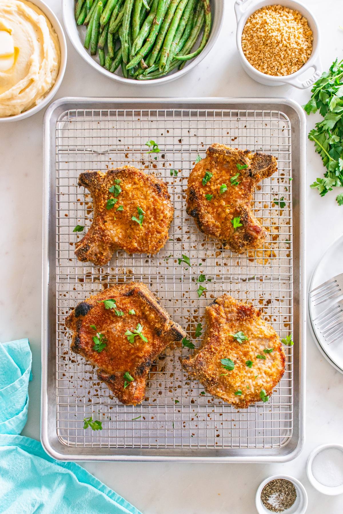 Four breaded pork chops on a wire rack with mashed potatoes, green beans, and seasonings on the side.