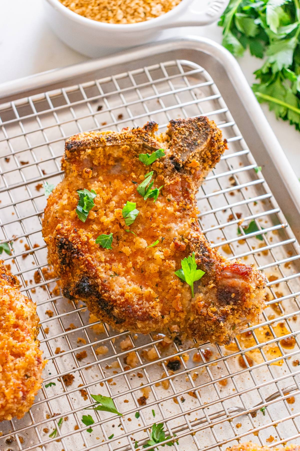 Breaded baked pork chop with parsley garnish on a cooling rack, with fresh herbs and breadcrumbs nearby.