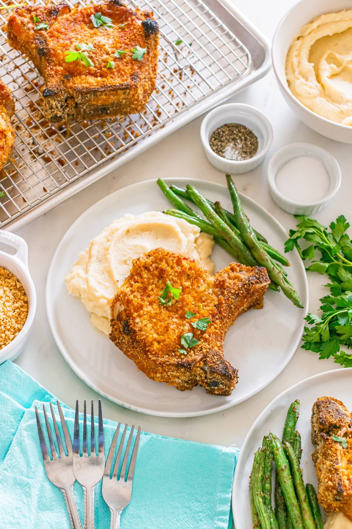 A breaded pork chop with mashed potatoes and green beans on a plate, surrounded by herbs and seasonings.
