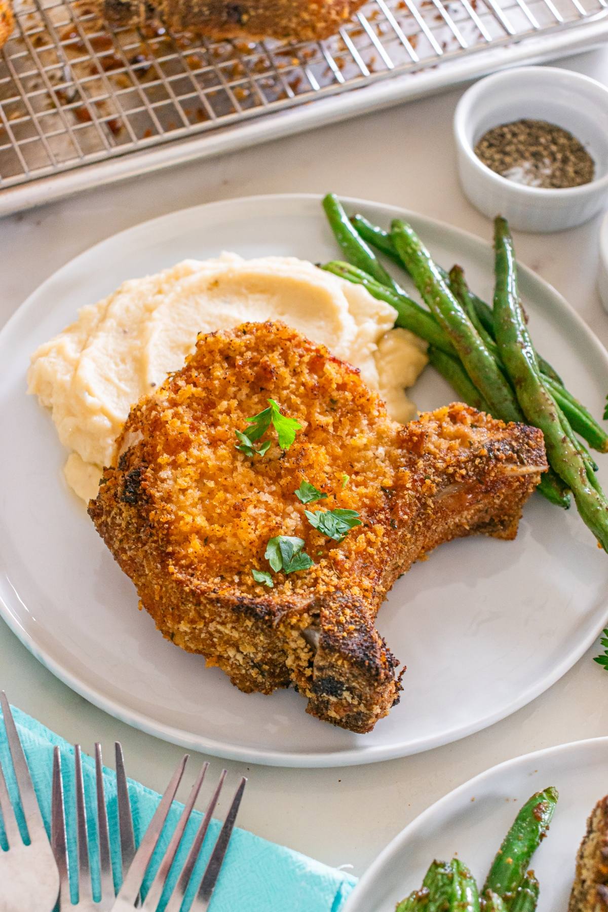 Breaded pork chop with mashed potatoes and green beans on a white plate, garnished with parsley.
