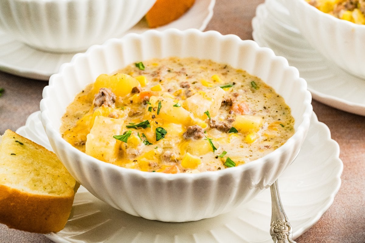 Slow Cooker Cheeseburger Soup: Creamy soup with ground meat, potatoes, and vegetables in a white bowl, served with a slice of garlic bread.