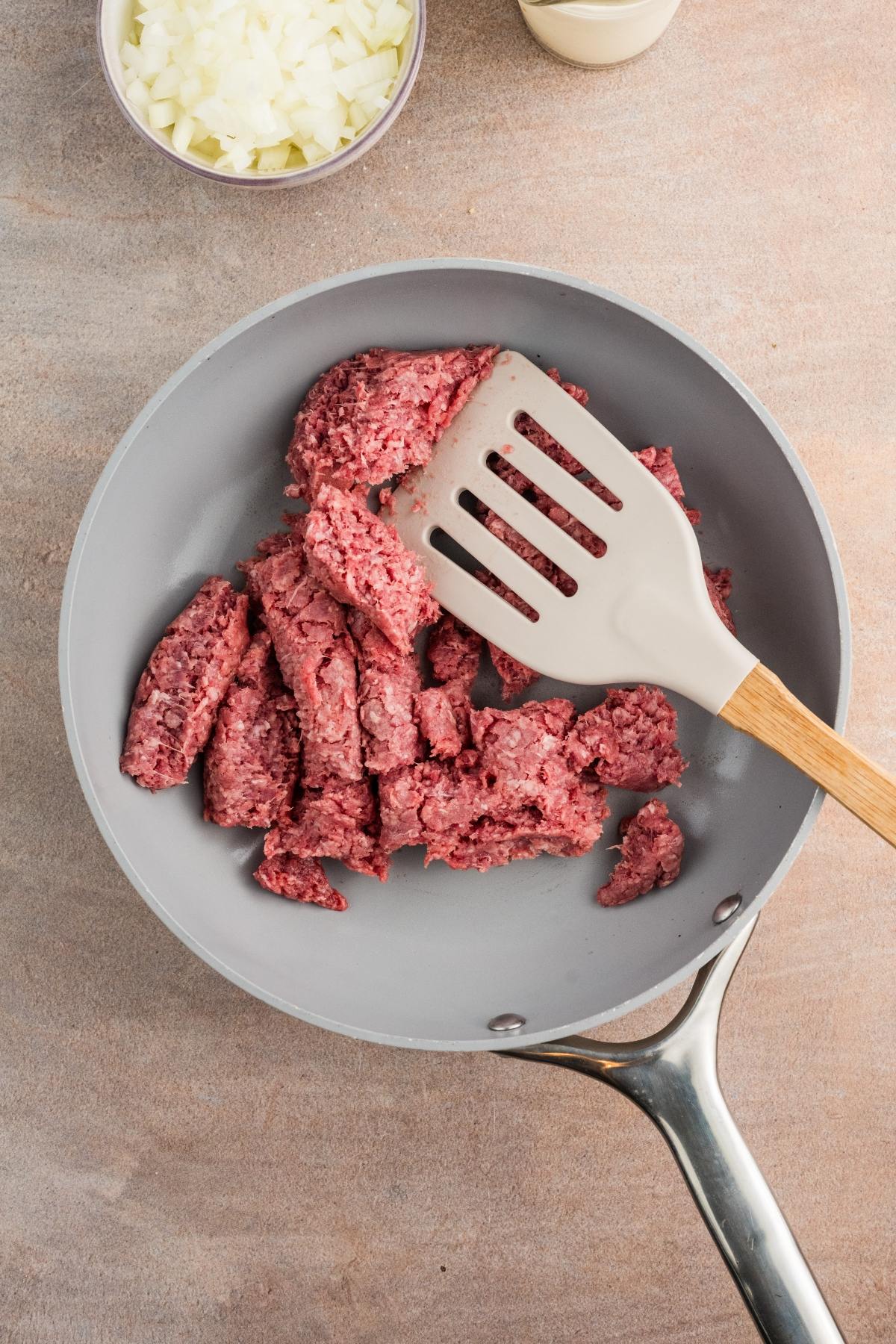Raw ground beef being browned in a skillet with a spatula, with chopped onions in a bowl nearby.