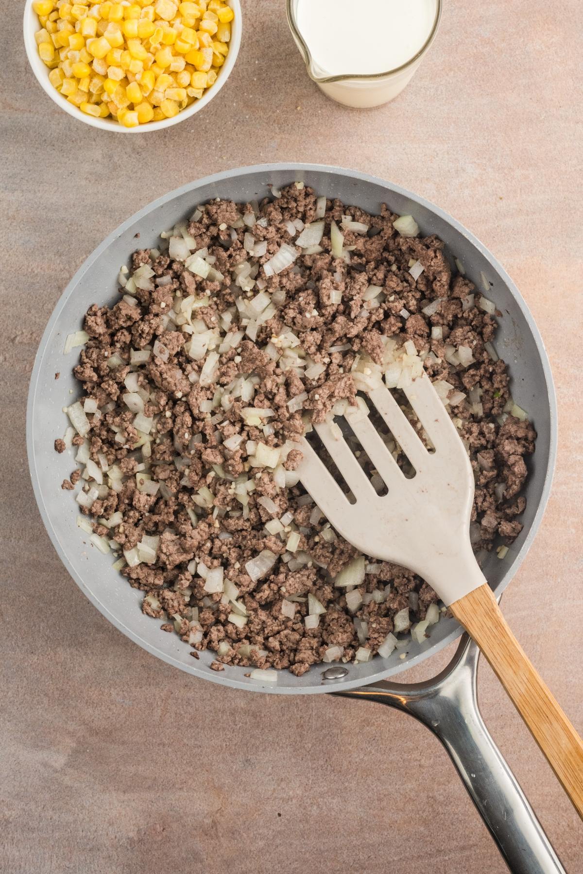 A skillet with cooked ground beef and chopped onions, a wooden spatula, corn, and a cup of milk nearby.