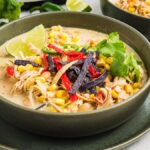 A bowl of creamy Slow Cooker Green Chile Chicken Soup topped with corn, tortilla strips, cilantro, and a lime wedge.