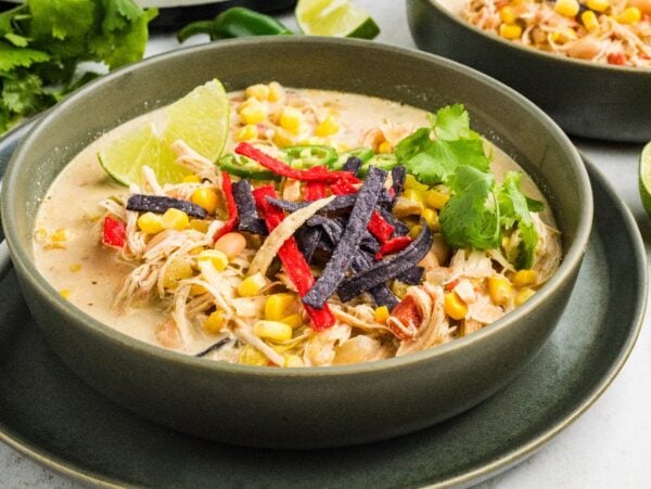 A bowl of creamy Slow Cooker Green Chile Chicken Soup topped with corn, tortilla strips, cilantro, and a lime wedge.