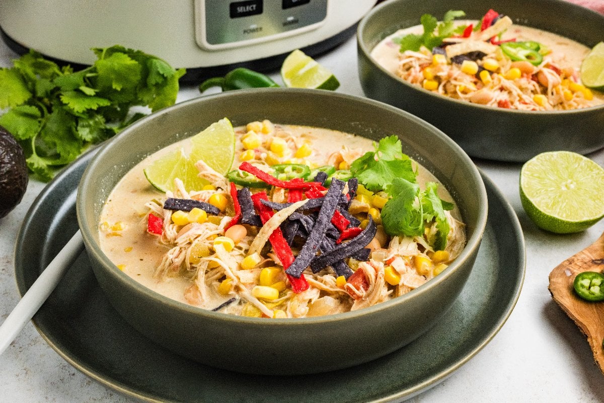 A bowl of creamy Slow Cooker Green Chile Chicken Soup with corn, cilantro, lime, and tortilla strips, garnished with herbs.