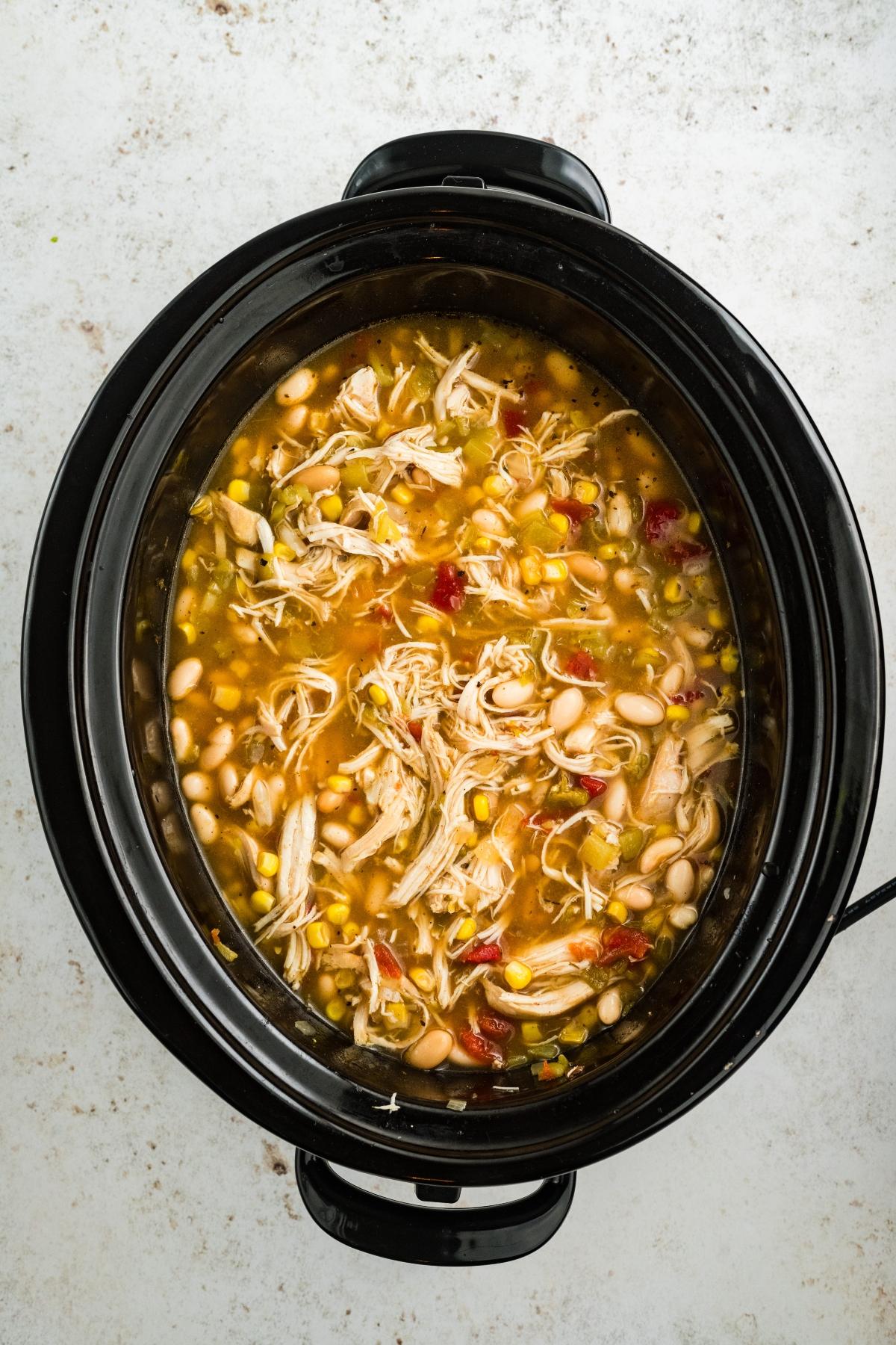 Slow cooker filled with shredded chicken, beans, corn, and vegetables in a broth, viewed from above.
