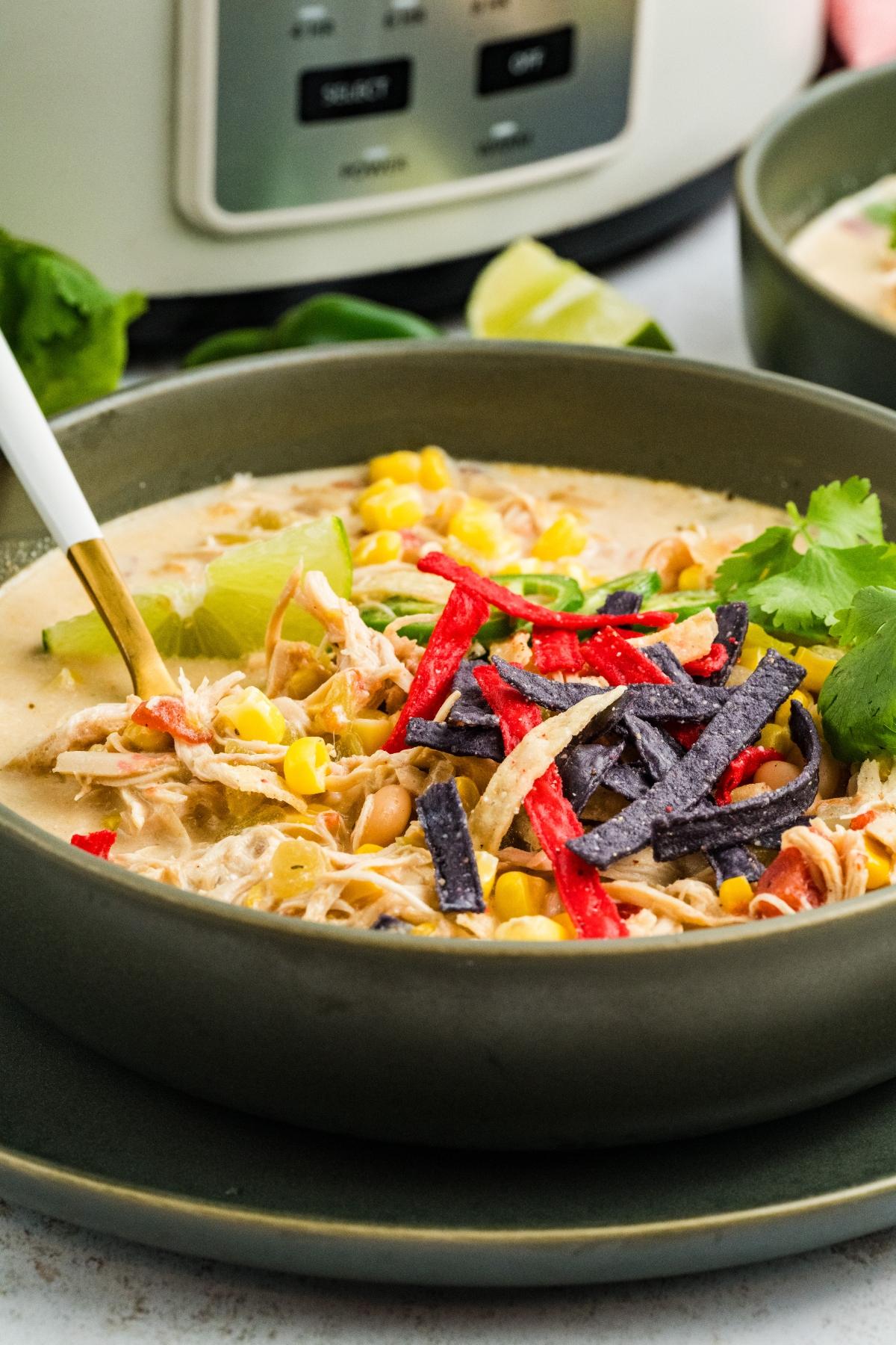 A bowl of creamy slow cooker green chile chicken soup topped with corn, tortilla strips, and cilantro, with a slow cooker in the background.