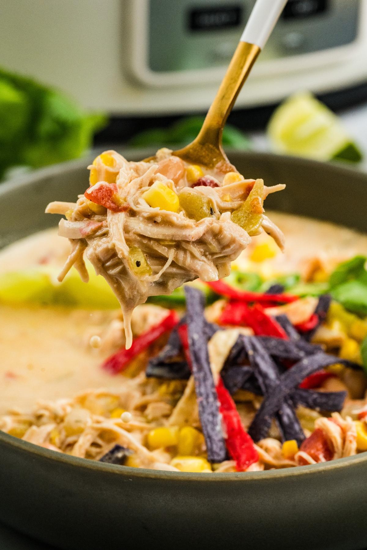 A spoonful of creamy chicken soup with corn, peppers, and tortilla strips above a bowl of soup.