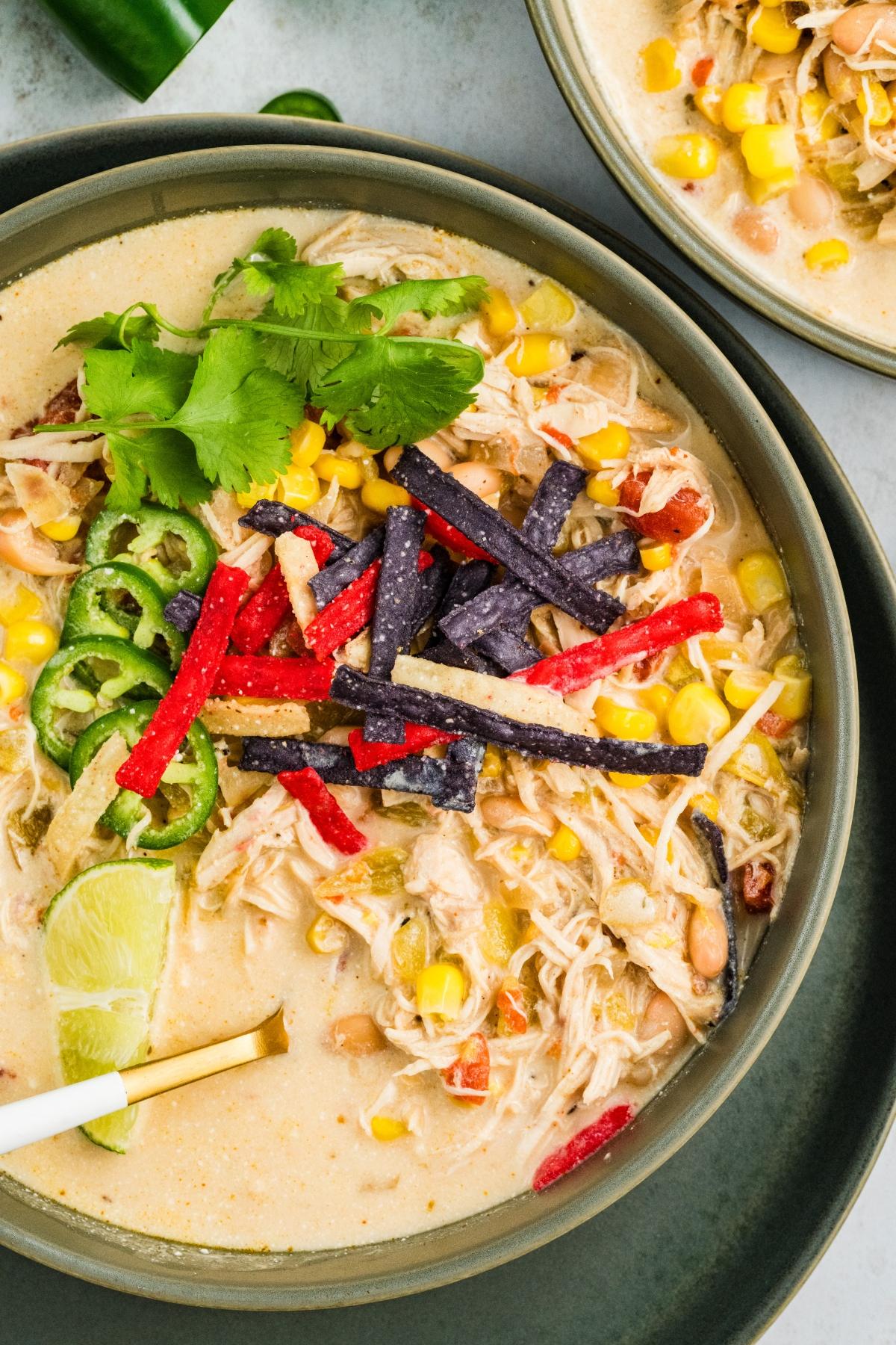 A bowl of creamy  slow cooker green chile  chicken soup topped with tortilla strips, corn, jalape&ntilde;os, cilantro, and a lime wedge.