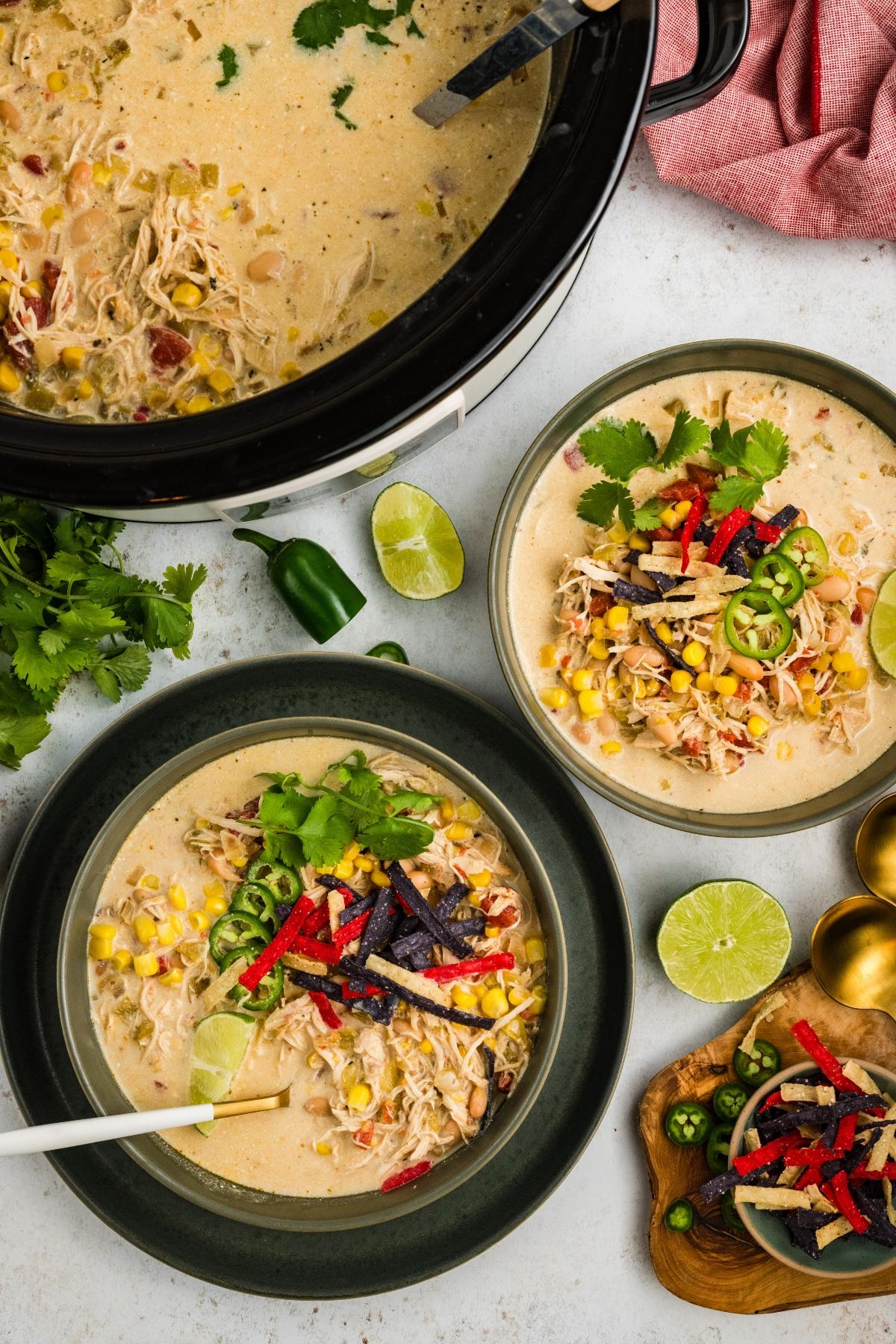 Two bowls of creamy chicken soup with corn and tortilla strips, beside a pot of soup, lime, and fresh cilantro.