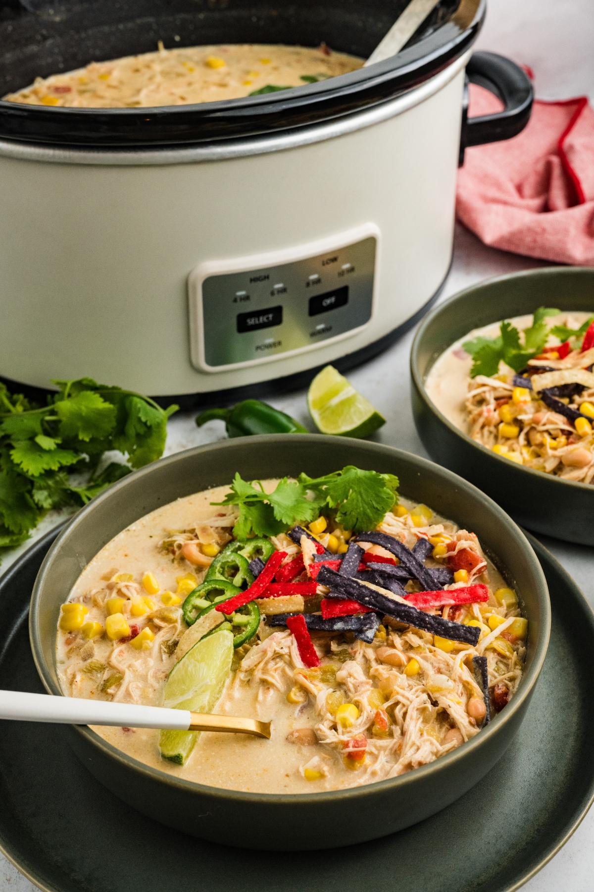 Two bowls of creamy  slow cooker green chile  chicken soup with corn, tortilla strips, jalape&ntilde;o, and lime, next to a slow cooker.