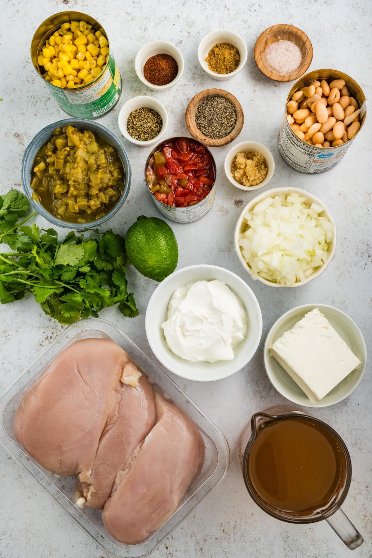 Ingredients for chicken chili laid out: raw chicken, canned beans, corn, spices, lime, herbs, broth, onions, and cheese.