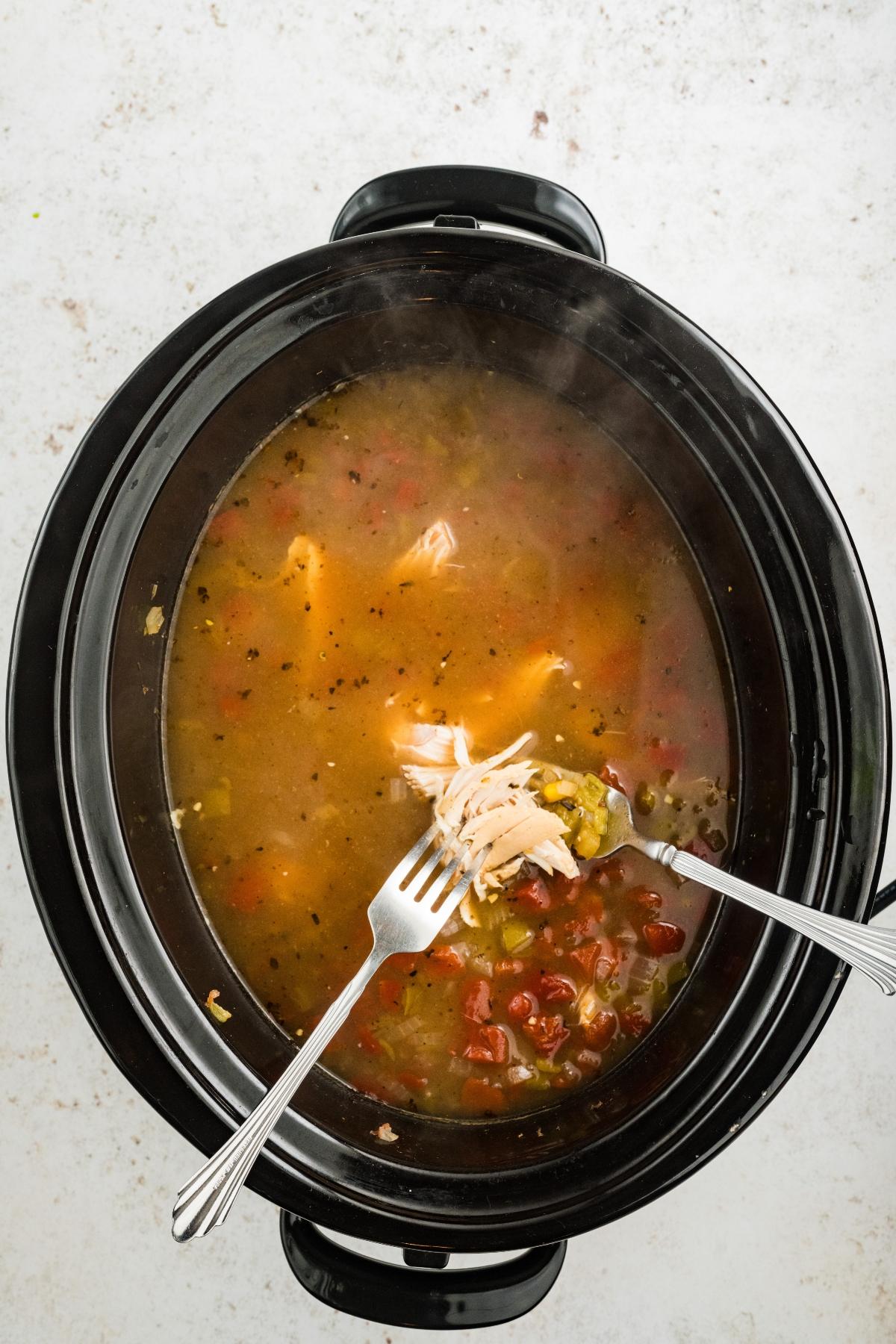 Two forks shredding chicken in a slow cooker filled with broth and vegetables.