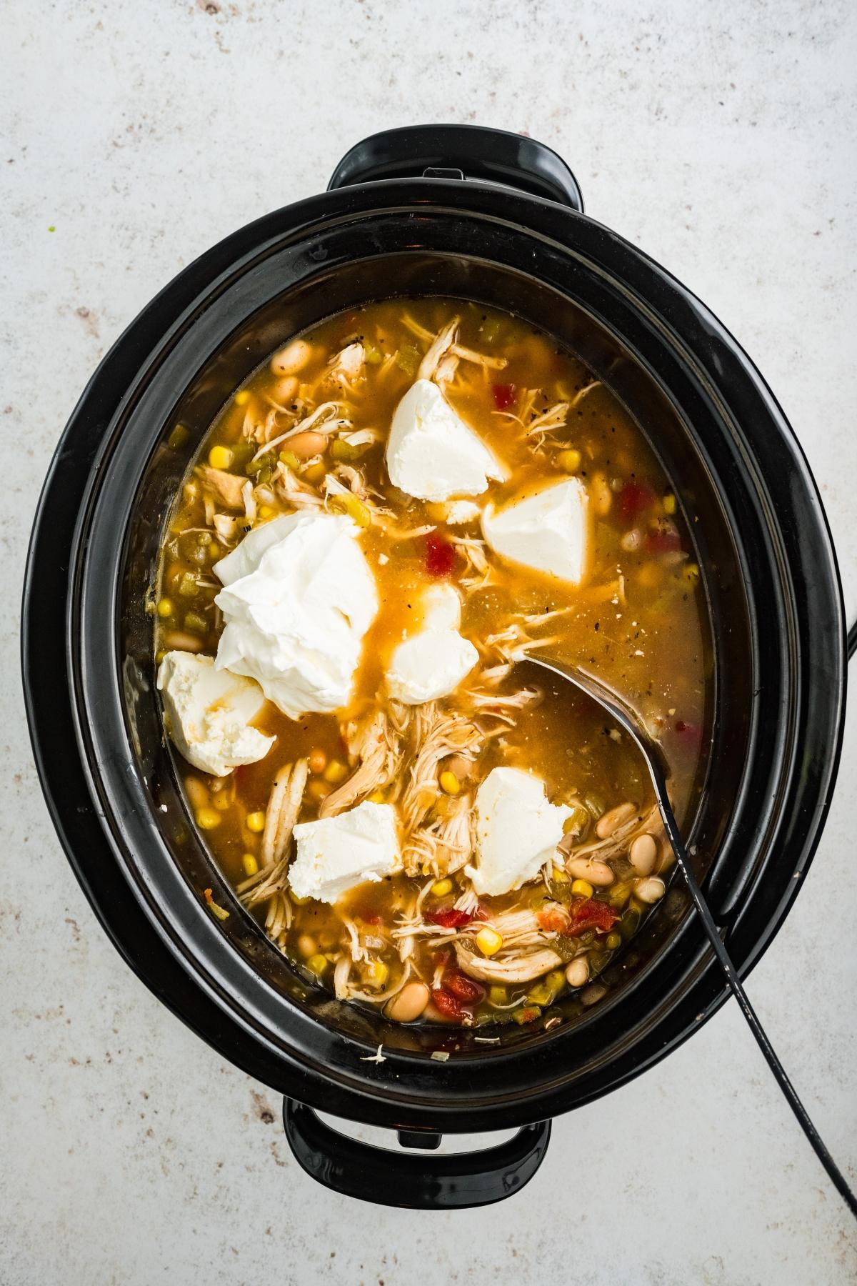 A slow cooker filled with white chicken chili, topped with cream cheese, and a spoon inside.