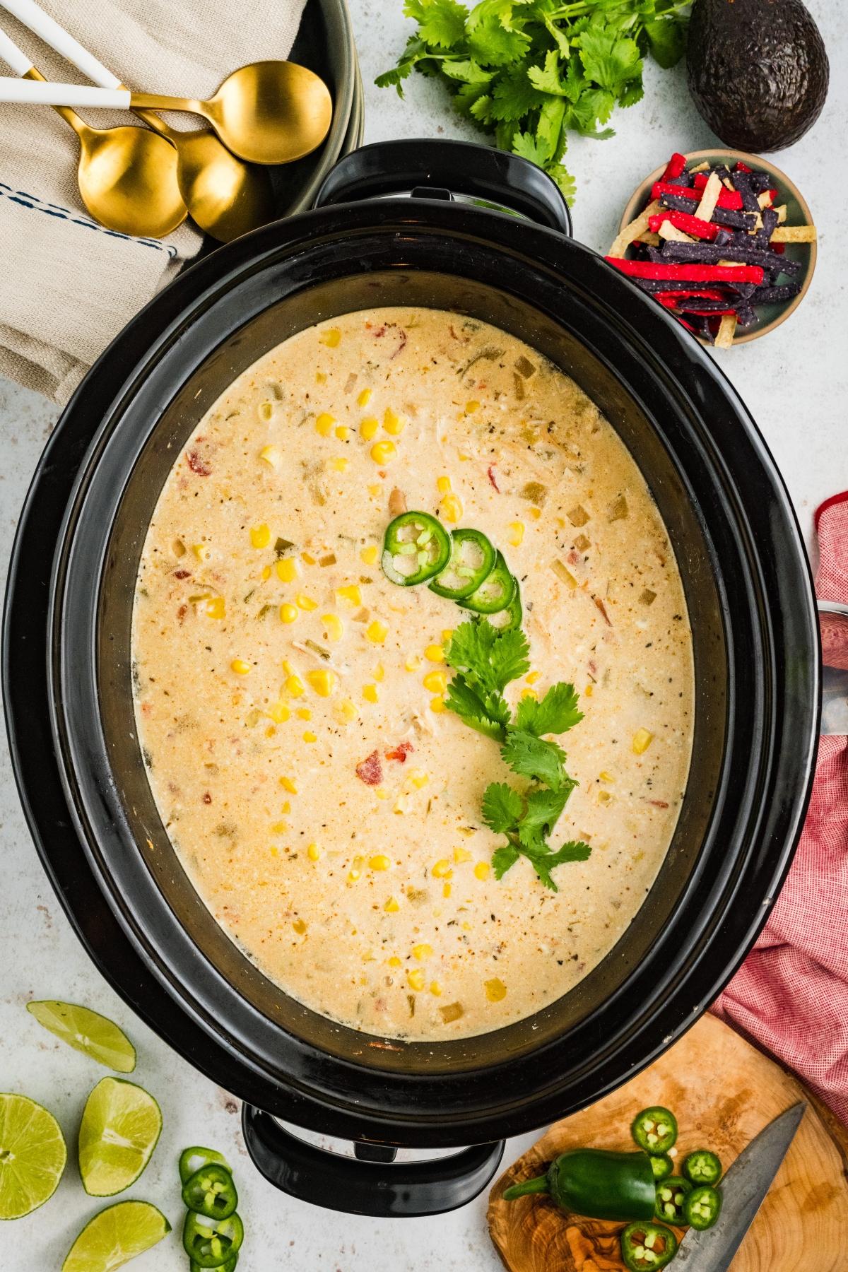 Creamy soup with corn and sliced jalape&ntilde;os in a slow cooker, garnished with cilantro, surrounded by toppings.
