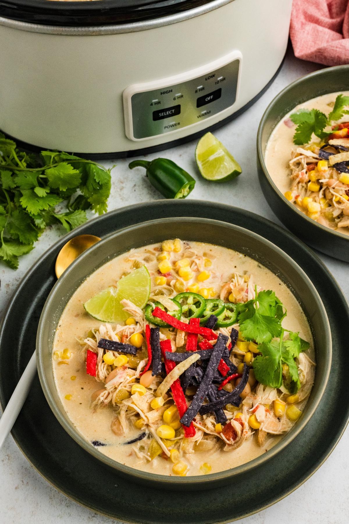 Bowl of creamy  slow cooker green chile  chicken soup topped with tortilla strips, cilantro, lime, and jalape&ntilde;o, with a slow cooker nearby.