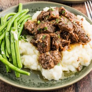 Plate with mashed potatoes topped with savory Slow Cooker Steak Bites, served with green beans on the side, on a wooden table.