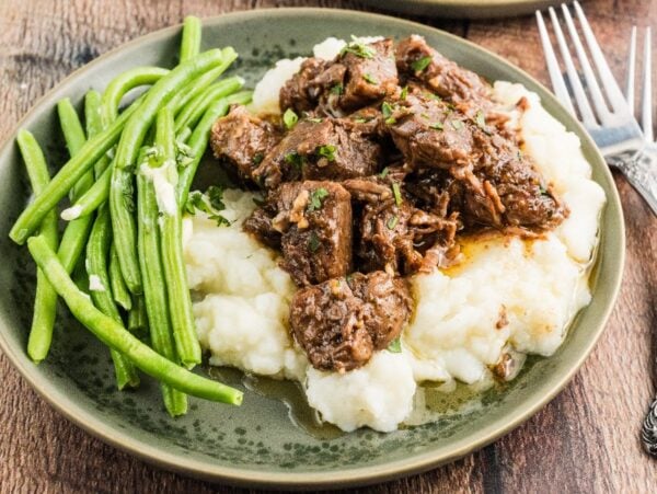 Plate with mashed potatoes topped with savory Slow Cooker Steak Bites, served with green beans on the side, on a wooden table.