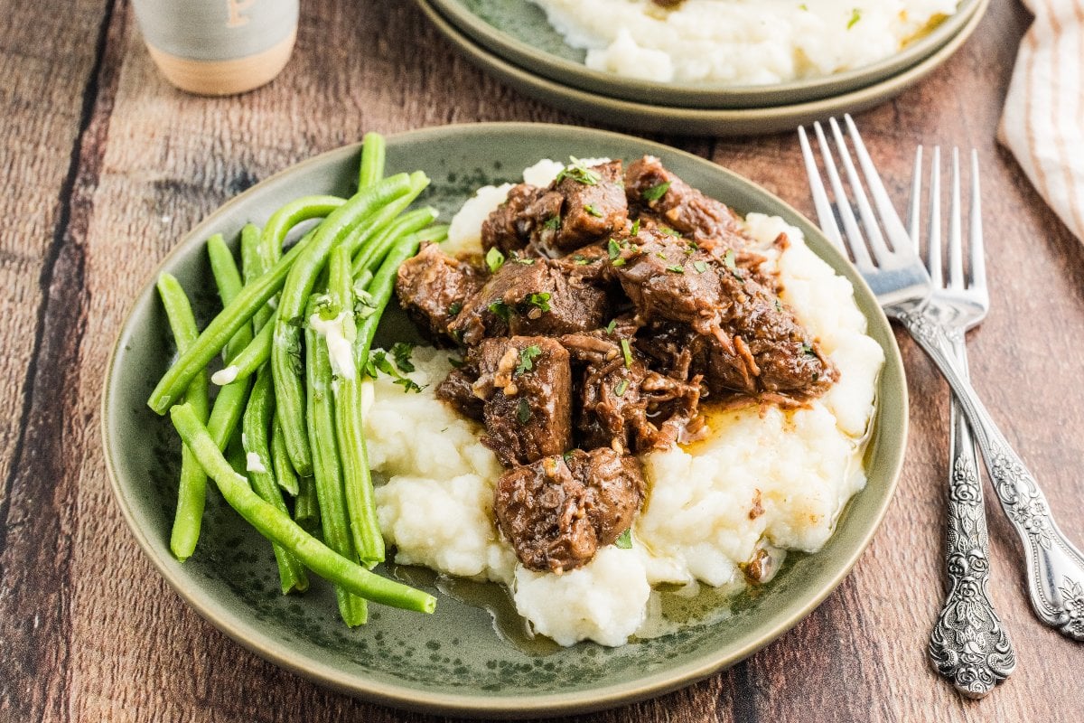 Plate of mashed potatoes topped with savory Slow Cooker Steak Bites, served with green beans on the side.