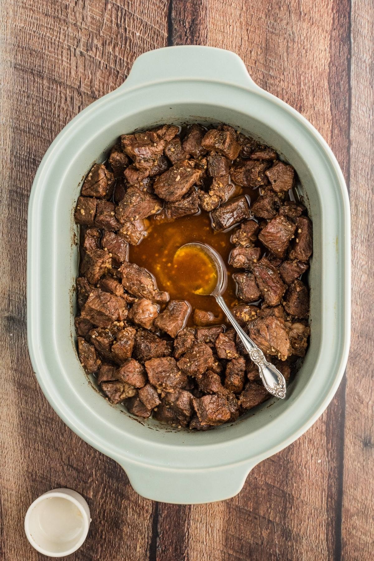 Chunks of cooked beef with broth in a slow cooker, with a ladle, on a wooden table.