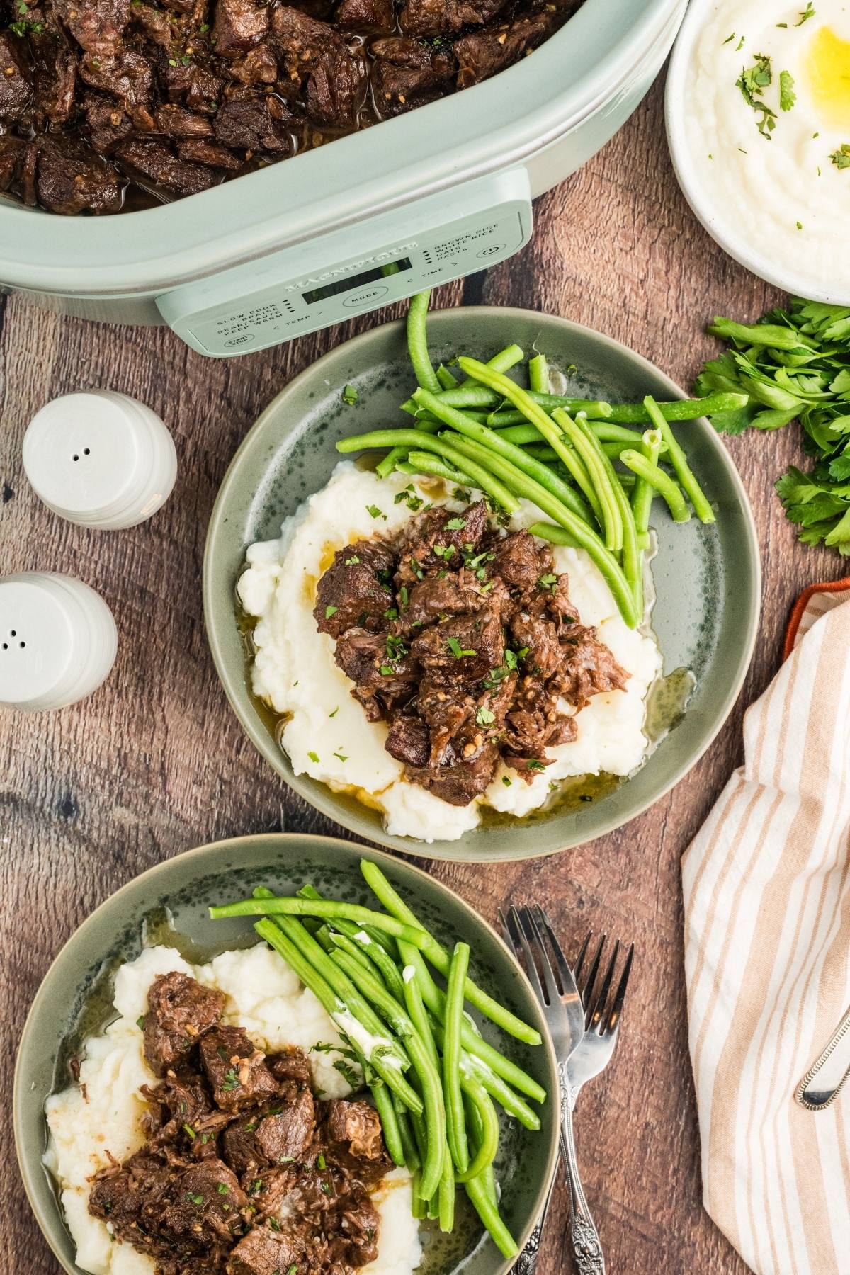 Two plates of beef stew over mashed potatoes with green beans, next to a slow cooker and salt and pepper shakers.