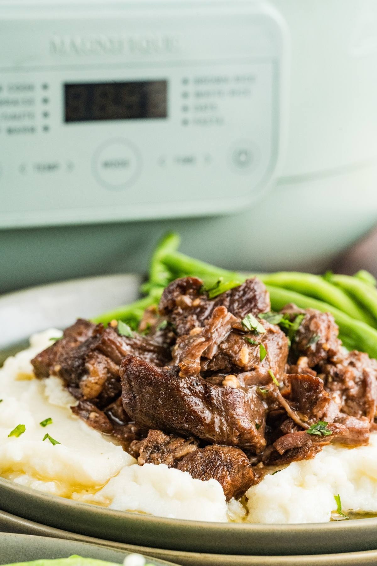 slow cooker steak bites served over mashed potatoes on a plate, with green beans in the background and a slow cooker behind.