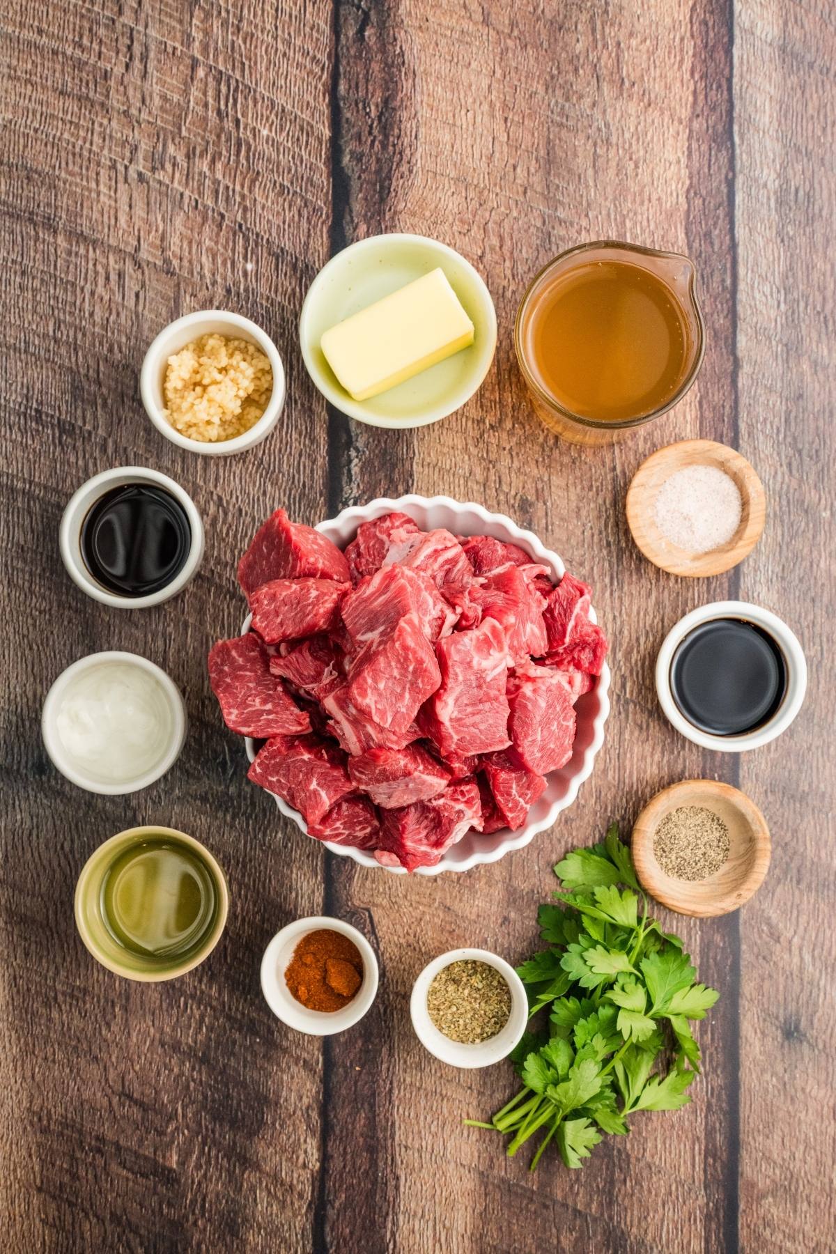 A bowl of raw beef cubes surrounded by small bowls of spices, butter, broth, and fresh herbs on a wooden surface.