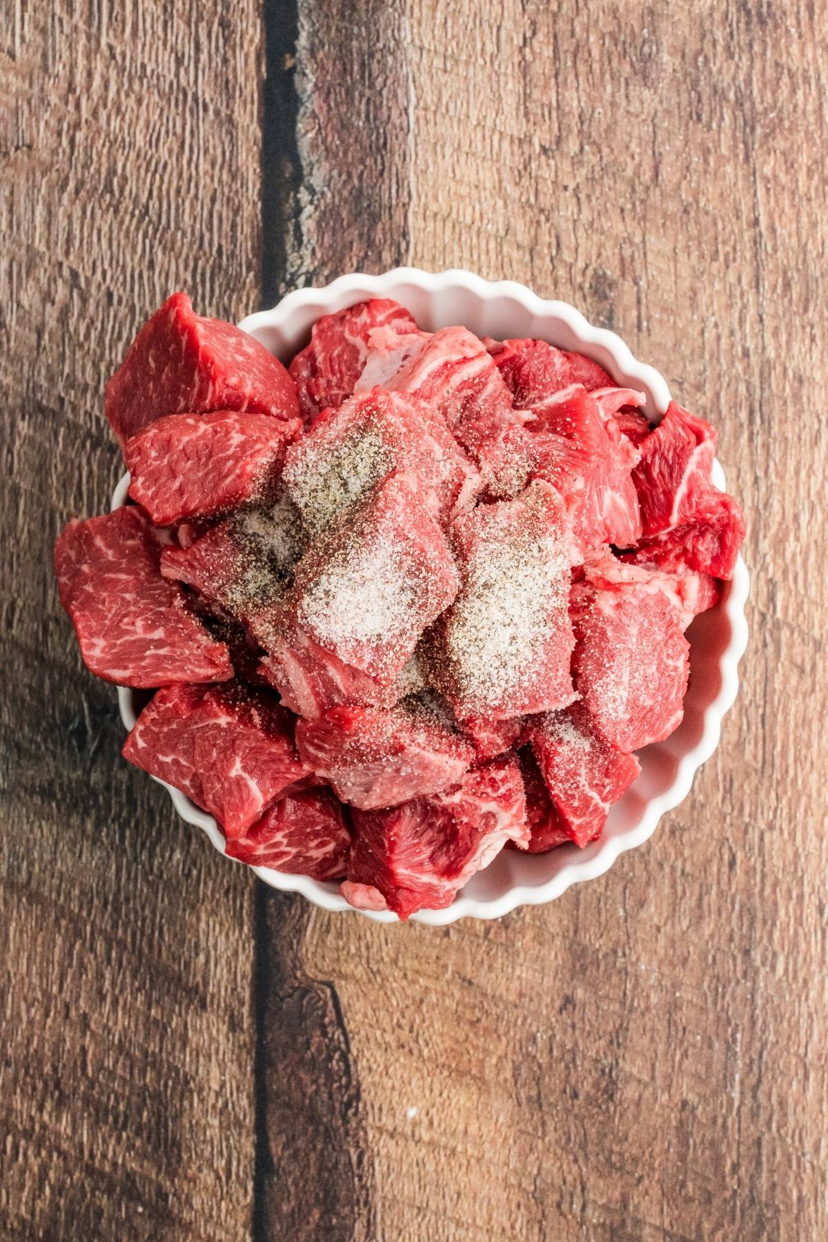 Bowl of raw beef chunks sprinkled with salt and pepper on a wooden surface.