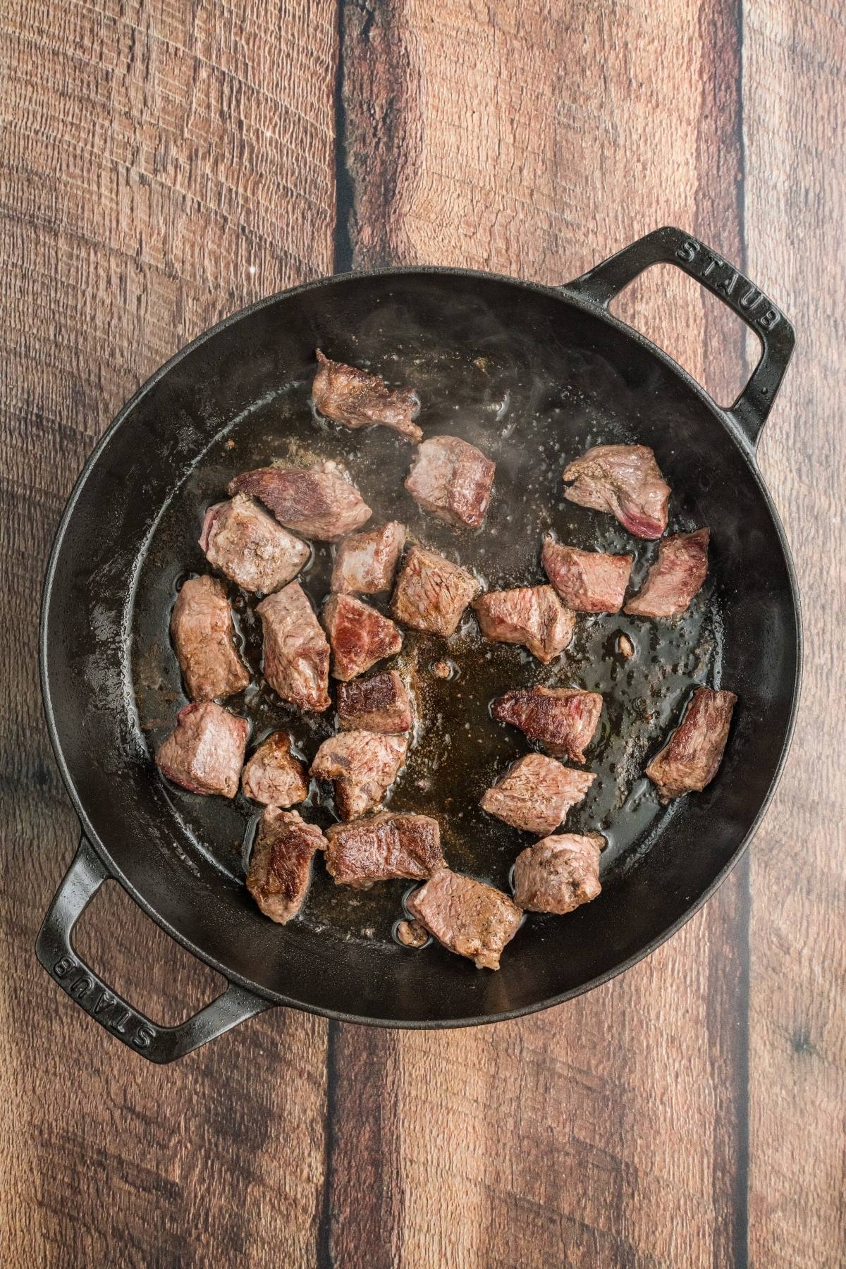 Chunks of browned beef cooking in a black cast iron skillet on a wooden surface.