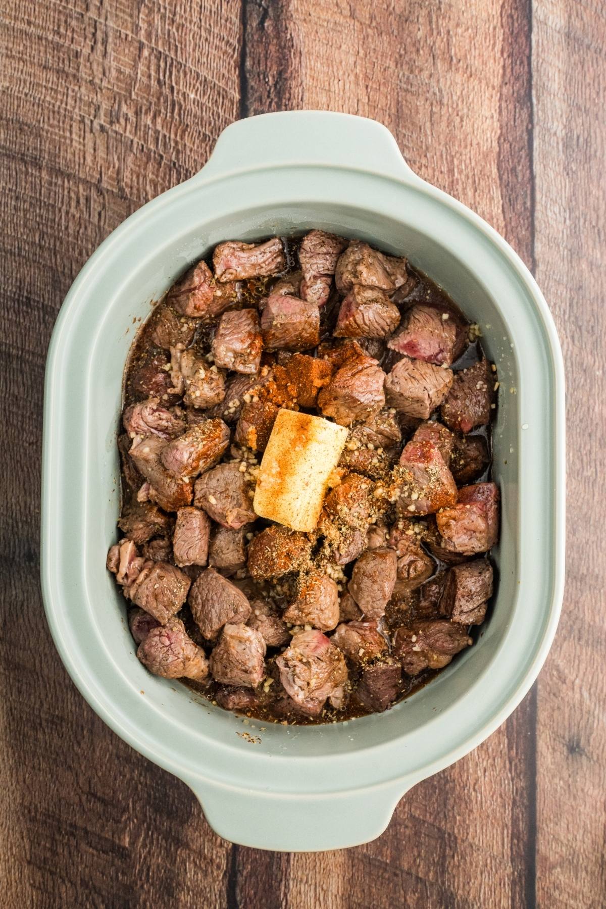 Chunks of raw beef and seasonings with a pat of butter in a slow cooker on a wooden surface.